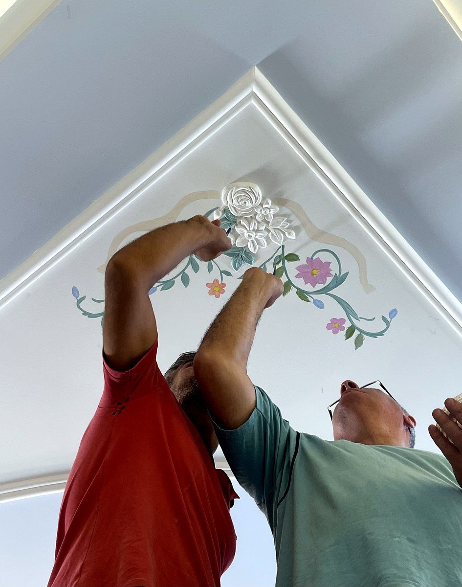 Two artisans painting an ornamental ceiling cornice